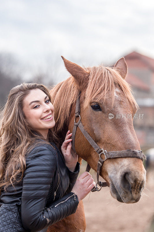 美丽的年轻女子用她的一只手牵着她棕色的马的缰绳，用另一只手抚摸/抚摸着马。她在镜头前很可爱。